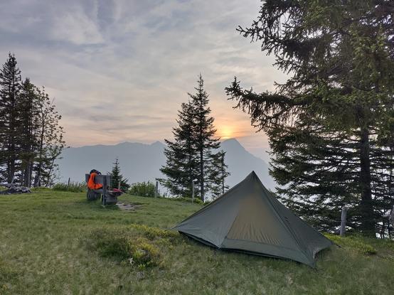 A green tent next to a bench with some gear. In the back a mountain with the sunset just above it.