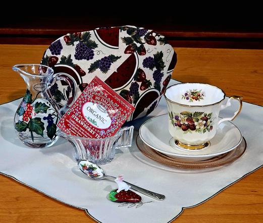 Tea tableau celebrating summer fruits with linen, china, silverware patterned in fruits, primarily berries, and even a rock painted to look like a strawberry. The tea is Strawberry black tea from St Dalfour. There is a wee white glass rabbit perched on the strawberry rock.