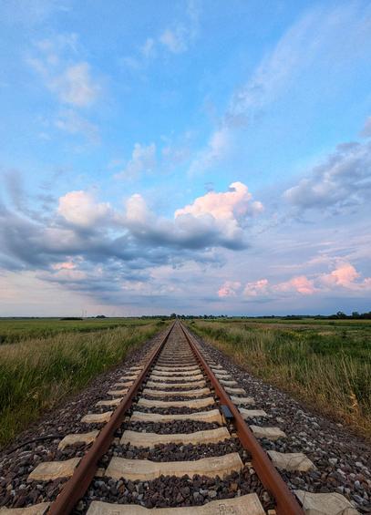 A low-angle shot of a single railway track stretching into the distance under a vast sky with scattered clouds. The track, made of rusty rails on wooden sleepers and gravel ballast, bisects a flat, green field that extends to the horizon on both sides. The sky is predominantly blue with patches of white and pink-tinged clouds, suggesting sunset. The perspective makes the tracks appear to converge in the far distance, creating a strong sense of depth.