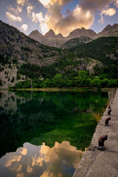 A vertical photograph showing a mountain landscape at sunset. In the foreground, a lake can be seen, and in the background, the Aragonese Pyrenees mountains, with the sun already behind them.