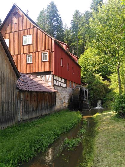 Die Mühle. Ein großes Haus mit roter Holzfassade, unten ist eine Steinwand und zwischen der Holzfassade und der Steinwand ist auch etwas Fachwerk zu sehen. Rechts an der Mühle fließt ein Bach entlang, er kommt über einen kleinen Wasserfall aus dem Wald. Dort ist auch ein Mühlrad aus Holz an der Steinmauer angebracht.