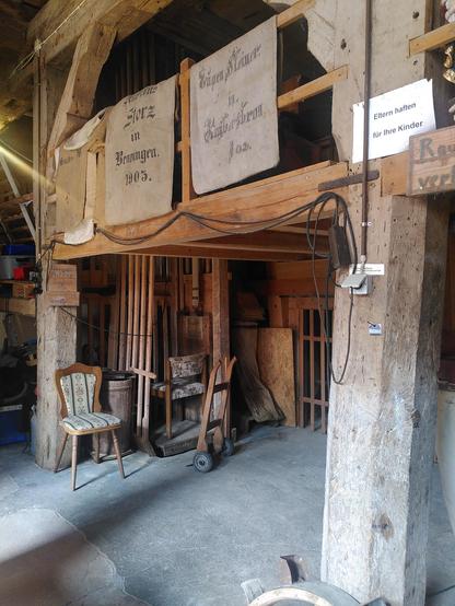 Untere Etage der Mühle. Viele Teile sind aus Holz gefertigt. Auf dem Geländer der oberen Etage hängen leere alte Leinensäcke. Es gibt einen Fahrstuhl, in die eine sitzende Person passen könnte. Er sieht aus wie eine aufgestellte Holzkiste. Darin steht ein gepolsterter altmodischer Stuhl, aber hauptsächlich wurden früher wahrscheinlich Säcke darin transportiert. Vor dem Fahrstuhl steht ein Sackkarren.