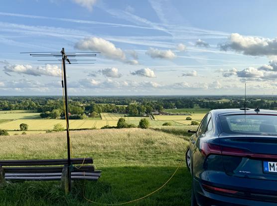 Das Bild zeigt eine idyllische Landschaft mit einem weiten Blick über Felder und Wälder. Im Vordergrund steht ein dunkelblaues Elektroauto (Audi e-tron), das an eine gelbe Leitung  angeschlossen ist, die zu einem kleinen Antennenmast führt. Neben dem Auto steht eine Parkbank. Der Mast ist an der Parkbank angebunden. Der Himmel ist leicht bewölkt, die Sonne scheint. Das Bild vermittelt ein Gefühl von Ruhe und Freiheit in der Natur.