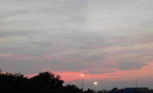 Un cielo al tramonto con tonalità soffuse di rosa, arancione e viola che si mescolano tra le nuvole. In basso, si vede la silhouette scura degli alberi e qualche edificio industriale sulla destra.