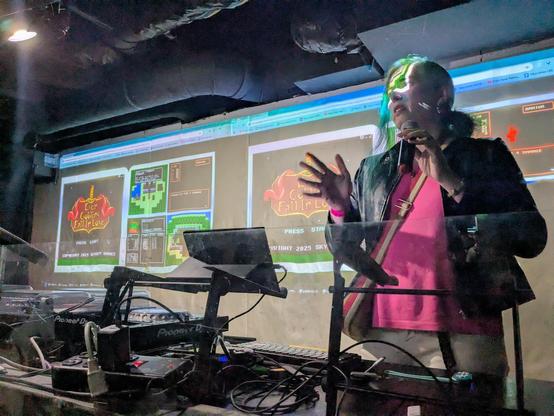 Robbie Dieterich, from Skyboy Games, doing a talk about her new NES JRPG, Even Goblins Fall In Love.
She is speaking from a DJ booth, wearing a leather jacket and a pink shirt. Colors from the projector wash across her face.