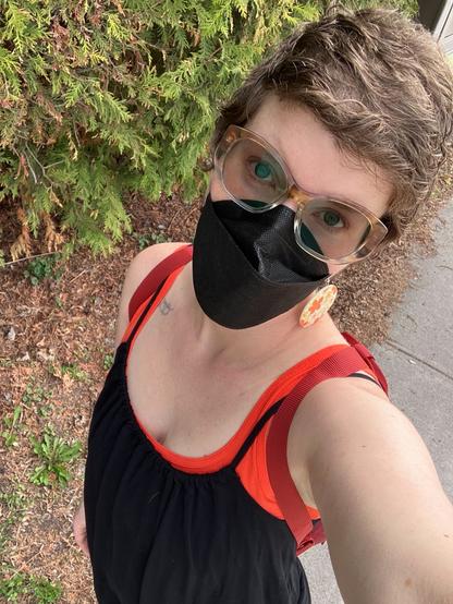 A middle aged white woman wearing a black n95 mask and sunglasses. She is dressed in a sundress with a backpack.