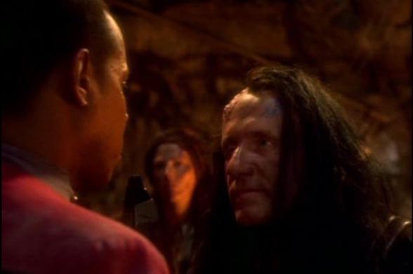 A tense confrontation between two male characters, one with short hair in a uniform and the other with long, dark hair and a rugged appearance, set against a dimly lit, rocky backdrop. Another figure is partially visible in the background.
