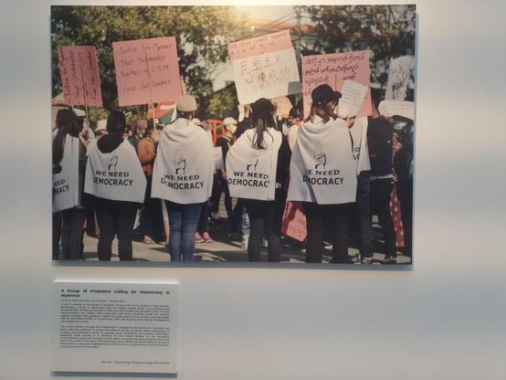 Photo of a group of protestors for democracy in Myanmar, showing people from the back wearing white capes that read "we need democracy".