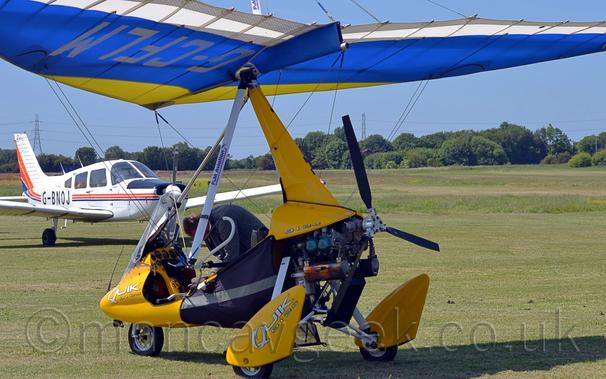 Rear side view of a single-engined microlight parked on grass facing to the left.
The plane has a yellow fuselage with black fabric covers under the fuselage, and an exposed engine at the rear.
The fuselage is suspended under a yellow, blue, and white fabric covered wing, with the registration "G-CFLM" underneath in silver.
A man wearing a grey coat can be seen leaning over the side of the cockpit from the other side.
A white, slightly bigger light aircraft can be seen in the background on the left of the frame, the rest of frame being green grass leading up to trees in the distance, under gorgeous blue sky.