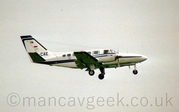 Side view of a twin engined BizProp flying from left to right at a low altitude with undercarriage lowered and flaps extended from the rear of the wings, suggesting it is about to land.
The plane is mostly white, with blue stripes running along the body and over the engines, with the last 3 letters of the registration "-CAE" visible at the bottom of the tail, under a small Slovenian flag.
Flat white sky fills the rest of the frame.