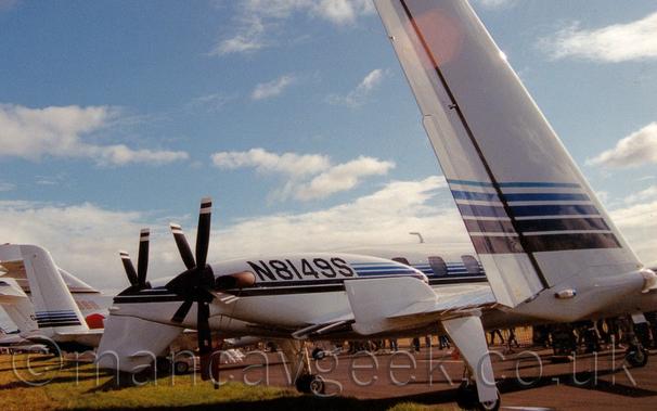 Close-up of the twin tails of a twin engined BizProp parked on bla
ck tarmac facing to the right.
The plane is mostly white, with a series of light and dark blue stripes running along the body and over the engines, which are mounted on the rear of the wings, facing backwards.
The black registration "N8149S" is painted on the sides of the engine cowlings.
As well as the twin tails on the ends of the wings, there is a small ventral tail under the rear fuselage - whether that is for additional stability, or to stop the propellor blades hitting the ground on take off or landing is unknown, at least to me.
(It's probably a bit of both!)
The tails of several other planes can be seen in the background on the left, and the feet of people in the crowd can be seen under this plane.
Gorgeous blue sky with scattered fluffs of cloud fill the rest of the frame.