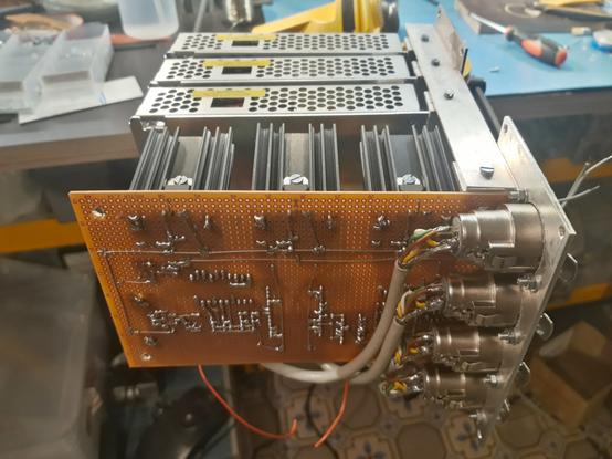 An electronic device on a workshop bench. Solid-looking connectors have wires that go behind an electronics board that is visible from the solder side. The board has big heatsinks and behind three shielded units are visible that are also part of the device.