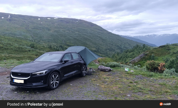 The image depicts a black Polestar car parked on a gravel surface in a natural setting. The car is positioned to the left of the frame, with a green tent partially covering it, suggesting a camping scenario. The background features a lush green hillside with patches of snow on the distant mountains, indicating a high-altitude or northern location. The sky is overcast, with clouds covering the majority of the visible sky. The ground is uneven with patches of grass and small rocks, typical of a mountainous terrain. In the foreground, there are some camping supplies, including a rolled-up sleeping bag and a white bag, placed on the ground. The image is credited to "u/jonsvi" and posted in the r/Polestar subreddit, as indicated by the text at the bottom of the image.