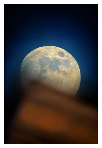 A waxing gibbous moon, nearly full, rises over a building rooftop in the extreme foreground. The blurred edge and corner of the rooftop partly obscure the moon’s lower third. The crater details are visible and the moon gives off a gently luminous glow against the black field of the sky.
