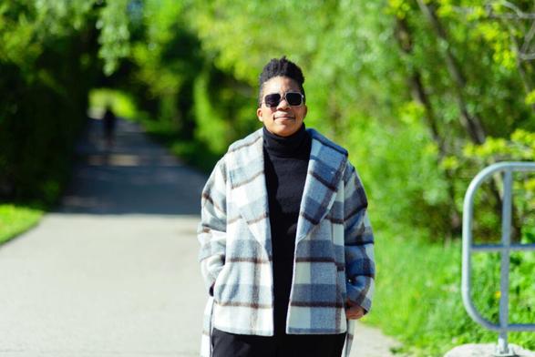 Photo taken outside on a sunny day of Sharl Smith, standing on a trail in the Sandrock Greenway wearing a plaid coat and square sunglasses.