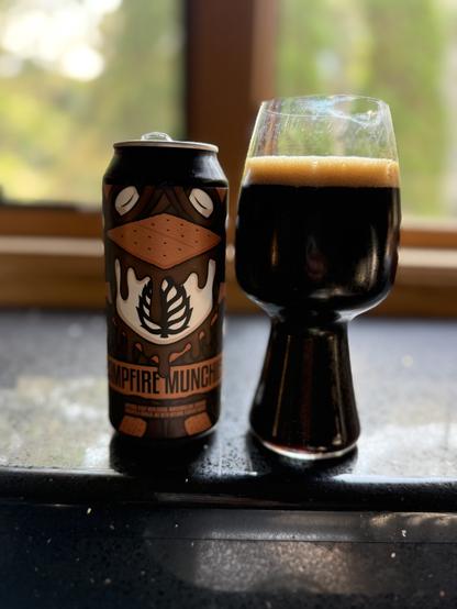 A stout glass full of dark beer with a tan head. The pint can is in shades of brown with a tan graham cracker and a white marshmallow covered in dark brown oozing chocolate