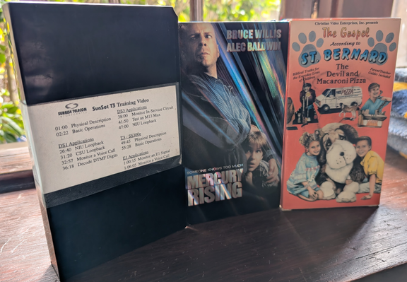 three tapes on a mantel. One is a training video for some inscrutable telecom device, one is MERCURY RISING, and one is The Gospel According To St Bernard: The Devil And Macaroni Pizza
