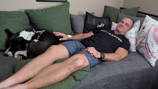 Man in shorts and T-Shirt laying on bed surrounded by pillows and medium sized black and white dog curled up next to his right side.