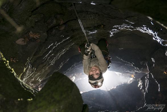 A photo, apparently so good it should be AI. The view is looking down into a pit, backlit from below which makes clear crisp highlights of the rock layers. Close to the entrance there is green highlighted due to the moss on the limestone. A rope hangs down, and a person in white helmet and shirt is attached to it, climbing. They lean back (towards the bottom of the image) to look up at the camera and smile. The dynamic lighting and clean lines and composition I guess is too good to be true. How sad is that?