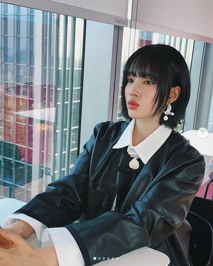 The image shows Kpop idol Yoon seated at a table with large windows in the background, offering a view of a cityscape with tall buildings. She is wearing a black leather jacket with a white collared shirt underneath, featuring a prominent white collar and cuffs. The jacket has a shiny texture, and the person is accessorized with large, white  pendant earrings that include a dove design. Her hair is short and black, with bangs framing the face. The setting appears to be a modern office or a similar professional environment, with natural light streaming in through the windows. The overall color scheme is neutral, with the black and white clothing contrasting against the light-colored table and the bright, reflective windows.

Provided by @altbot, edited by @jemmesedi