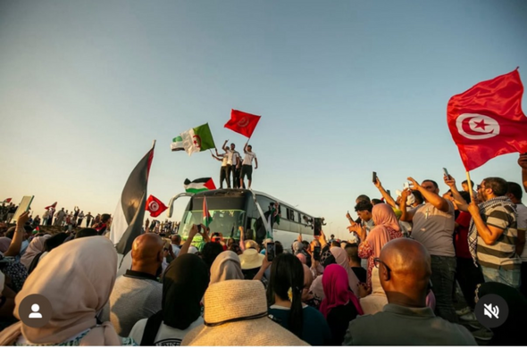 Photo of a large crowd of people surrounding a motorcoach, and some are on its roof. Many of the crowd are taking pictures with their phones, while others are waving flags of what appear to be North African countries and Palestine.