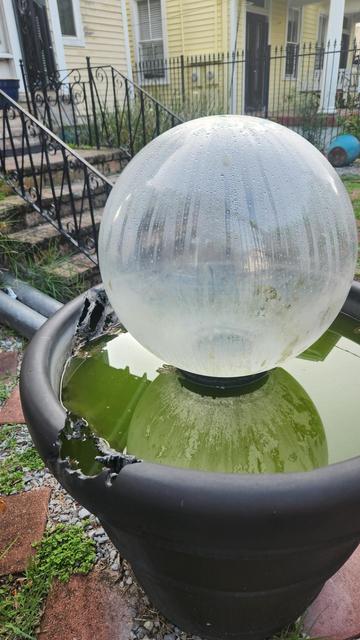 A large plastic pot full of water with a large glass gazing ball fountain in its center. The fountain is turned off, so the gazing ball is empty. The edge of the pot is melted in a large strip across one side, apparently from the sun being focused onto the edge as it traversed the sky.
