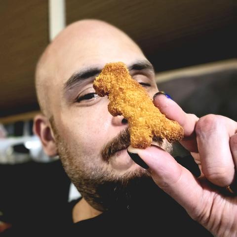 Matt B playfully holds a dinosaur-shaped chicken nugget in front of his face, partially covering one eye. The background is softly blurred, keeping the focus on the face and the nugget.