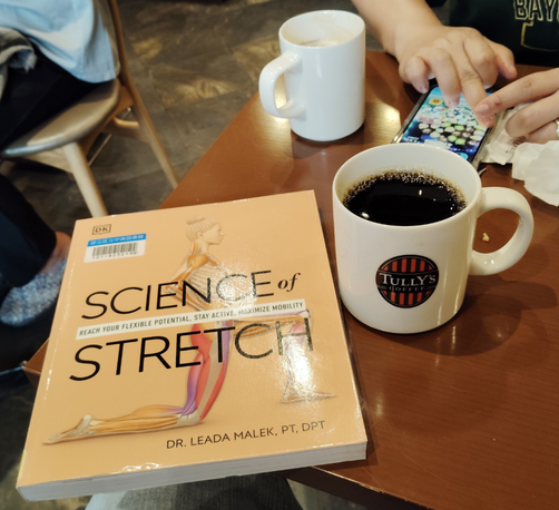 The wide tan paperback library book has an illustration of a woman doing a forward lunge stretch where her muscular system is shown. To the right is a white mug of black coffee with the Tully's logo on it. Behind it a woman is playing TsumTsum on her phone with a white mug with visible latte foam to the left