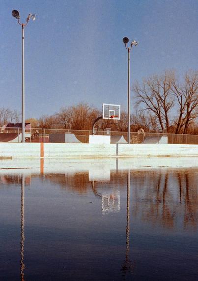 A flooded outdoor basketball court in a suburban park, with a hoop and light poles reflected in the still water. In the background, bare trees and a small playground hint at early spring. The image has a soft, vintage look typical of 35mm film.