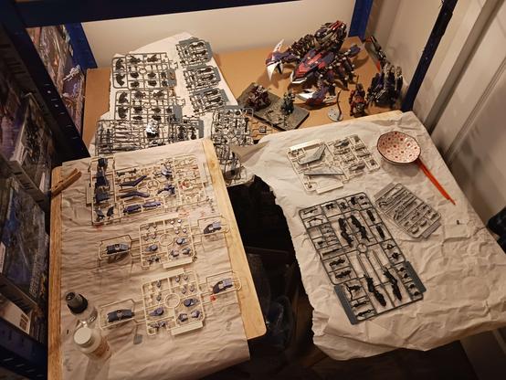A photograph of Amalgam's crafting "workshop" area, crunched in the corner of her bedroom blocking the closet door and sitting just in front of the display shelf. On the shelf are some build Zoids, tucked to the side out of the way (say hi to Ashley) and a drying paper with a bunch of black, finished sprues covering it. In front of and overlapping that are the two hobby tables, each with their own drying paper and three wolf sprues apiece, one set metallic blue, the other the gunmetal mixture (and the black sprue which got some of that for detailing some pieces). It's a cluttered mess and this isn't even all of it.