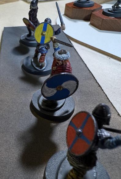 Four 28mm Vikings, each with roundshields. The nearest is red with a narrow blue cross on it, the next is swirly-quartered white and blue, the next is yellow with a vertical blue stripe down the middle. You can't really make out the back one.
