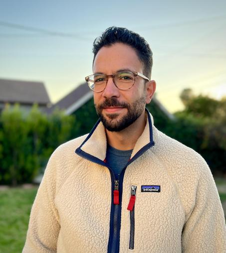 Head-and-shoulders color photo of writer Robert Nazar Arjoyan, a man with short, neatly trimmed hair, beard, and mustache. He wears a fleecy, cream-colored Patagonia zip pullover; a blue tee; and roundish tortoiseshell glasses. He stands on a green lawn, blurred green deciduous trees and gray house roofs behind him. He regards the camera with a small, amused smile.