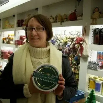 Waist-up color photo of Can Wiggins, a woman with straight brown hair worn in a beveled chin-length bob without bangs. She wears narrow-lensed glasses with dark frames, a cream-colored cable-knit sweater with a cowl neck, a dark winter jacket, and a cream-colored winter scarf. She stands in front of a wall of white shelves displaying gifts that appear to be for sale. She holds a tin marked CHRISTMAS PUDDING with a sly, or perhaps triumphant, little grin.