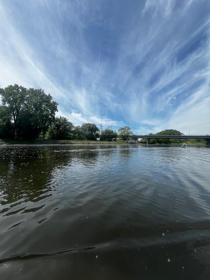 A brilliant, cloud-streaked sky over a wide expanse of river. An island with cottonwood trees is on the left - in the midground is a bridge across the river, underneath which a kayaker is headed.