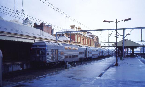 On a wet day, a silver and blue liveried double deck electric multiple unit train stands at a platform at a terminal station.  Another platform in the foreground is wet from a recent rain shower.  A large brick and stone station building is to the left of the platform on which the train is standing.
