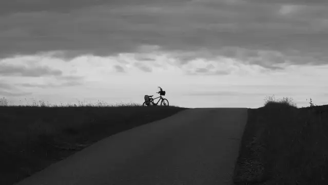 Schwarz weiß. Fahrradsilhouette auf einer Anhöhe. Oben graue Wolkendecke. In der Mitte weiße Wolken mit dem Fahrrad unten dunkler Boden
