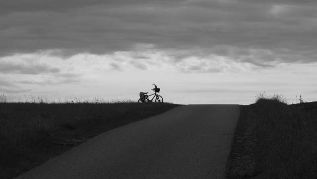 Schwarz weiß. Fahrradsilhouette auf einer Anhöhe. Oben graue Wolkendecke. In der Mitte weiße Wolken mit dem Fahrrad unten dunkler Boden