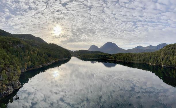 Wide landscape photo featuring a calm lake with reflections of the sky and surrounding forested hills. The sky is covered in a textured pattern of small clouds, and the sun peeks slightly from behind these clouds, creating a subtle glow. The panorama includes distinct silhouettes of rounded mountains in the background.