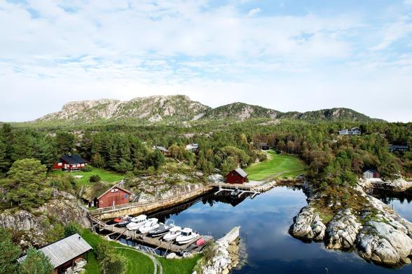 Aerial view of a scenic coastal landscape with lush greenery, several red and black wooden houses, and a marina filled with numerous small boats. The terrain features prominent rocky hills in the background, and a clear blue sky overhead. A narrow waterway reflects the surrounding elements, enhancing the serene quality of the setting.