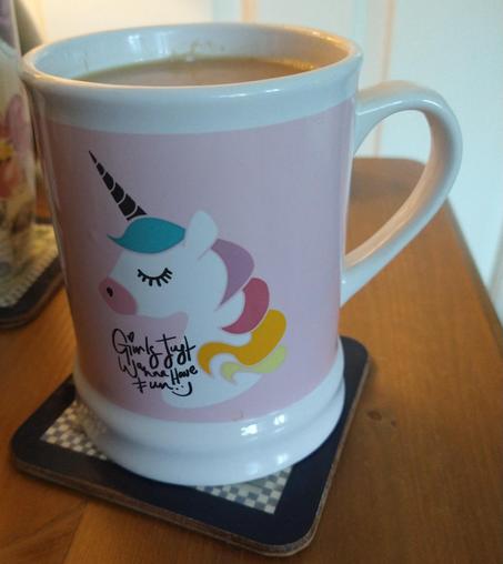 Tankard shaped mug of tea on a bedside cabinet. It has a pink background with a unicorn head with a multi coloured mane