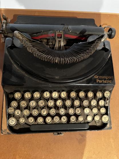 A typewriter. It’s black, with circular off-white keys. There’s a two-colour ribbon. It rests on a wooden base which forms part of its carrying case.
