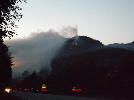 A wildfire at dusk on a steep slope producing a signifcant amount of with highway traffic in the lower forefront of the image.