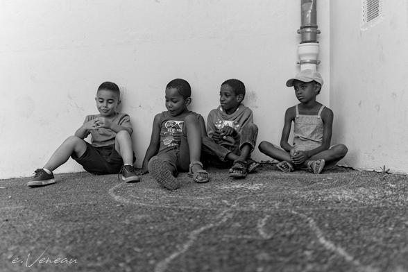 In a corner of the playground with chalk marks on the floor, four young pupils sit on the ground and seem to be talking to each other. The three boys on the left are smiling mischievously. The girl on the right looks puzzled or perhaps bored.