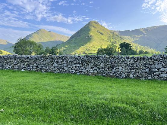 A flat patch of green grass, then a dry stone wall stretching right to left, and green-covered pointy hills in the background. Sun shining from the right so the hills cast shadows. Blue sky with flecks of cloud.