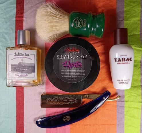 (Le Chat Mistral description)

The image showcases a collection of grooming items arranged on a striped cloth. At the top left, there's a bottle of aftershave labelled "Pure Lavender" from Chatillon Lux. Next to it, towards the center, is a round tin of shaving soap with a "Hipster" label from Smitten Soapery. To the right of the shaving soap, there's a bottle of Tabac Original Eau de Toilette.

Above the shaving soap, there's a green shaving brush with a wooden handle. Below the aftershave and shaving soap, there's a dark blue straight razor with intricate designs on the handle, branded H. Boker & Co. Tree Brand. The items are neatly laid out, suggesting a traditional shaving setup.