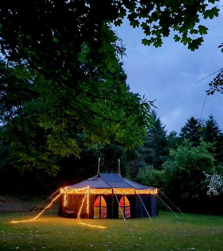 Auf dem Bild ist die Schutzhütte zu erkennen. Es ist eine Ovaljurte, die mit Lichterketten und 2 lichtdurchlässigen bunten Seitenplanen, die an Kirchenfenster erinnern sollen, einladend gestaltet wurde. Die Jurte steht auf einer Wiese, umringt von Bäumen. Das Bild wurde in der Abenddämmerung aufgenommen.