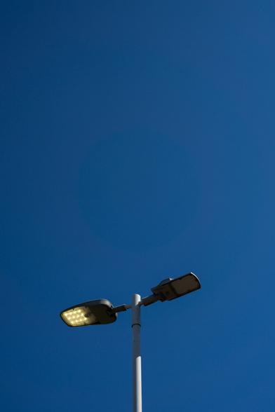 Photo of a clear deep blue sky. At the bottom is the top of a double-headed lamppost, the flat heads pointing away from each other, the one on the left lit, the other dark. Bright sunlight illuminates the right of the post, except where the lamp on that side casts a short shadow.