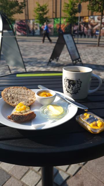 Café table outside with plate of a half eaten bun with orange jam, a case with yellow in-ears and coffee mug with a logo on.