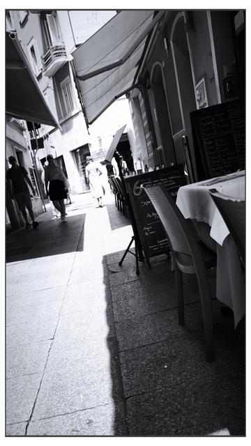 Fotografia in bianco e nero scattata lungo un vicolo urbano in una zona pedonale. Il lato destro è occupato dai tavolini di un ristorante, con una lavagna che mostra il menù del giorno. A sinistra, alcune persone camminano sotto gli ombrelloni e le tende dei negozi. La luce solare filtra dall’alto, creando un forte contrasto tra ombra e luce lungo il selciato.