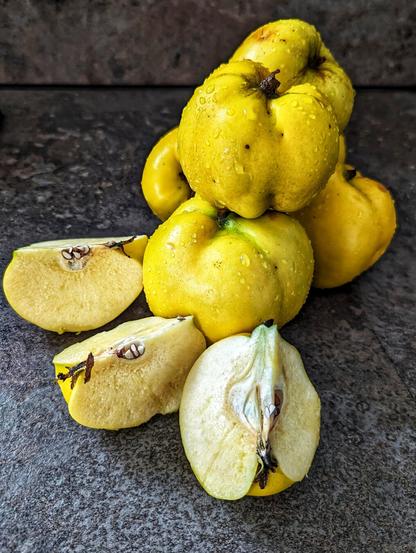 A close-up shot shows several yellow quinces, some whole and some cut, on a dark, mottled surface. The quinces appear to have water droplets on their skin. In the foreground, two wedge-shaped slices and one half of a quince reveal the light yellow flesh and dark seeds within. In the background, a pile of whole quinces is visible, some stacked on top of each other. The lighting highlights the texture of the fruit and the surface it rests on.